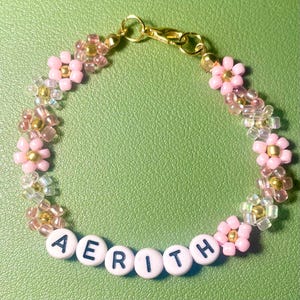 Puede incluir: Una pulsera de cuentas con detalles de flores rosas y el nombre "AERITH" escrito con cuentas de letras blancas. La pulsera tiene un cierre dorado y está sobre un fondo verde.