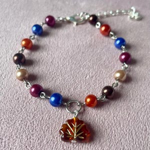 May include: A bracelet with a silver chain and clasp, adorned with multi-coloured faux pearls in shades of blue, orange, burgundy, and brown. It features an amber-coloured leaf charm.