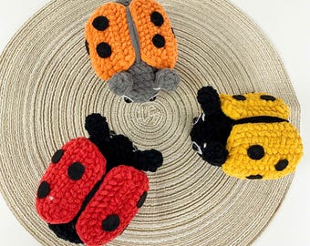 May include: Three handmade ladybug plush toys in orange, red, and yellow, each with black spots. The ladybugs are made of a soft, textured material and are arranged on a beige, circular woven mat.