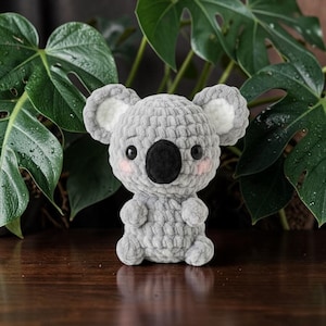 Puede incluir: Un koala de ganchillo gris hecho a mano con una nariz negra, ojos negros y mejillas rosadas. El koala tiene orejas grandes y redondas con detalles blancos en el interior. El peluche está sentado, mostrando sus pequeños brazos y piernas.