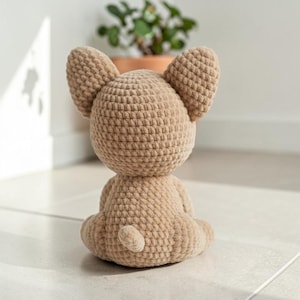 May include: A crocheted tan-colored dog figurine, viewed from the back, with large, upright ears and a small tail. The handmade dog is sitting on a light-colored surface. The dog is a soft, plush toy.