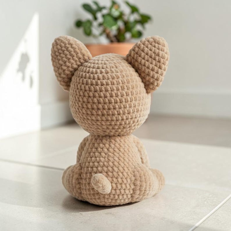 May include: A crocheted tan-colored dog figurine, viewed from the back, with large, upright ears and a small tail. The handmade dog is sitting on a light-colored surface. The dog is a soft, plush toy.