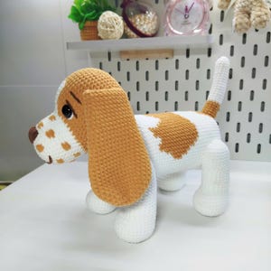 May include: A handmade crochet dog in a basset hound style. The dog is primarily white with brown and tan accents on the ears, spots, and back. The dog has a long, floppy ear and a short tail.