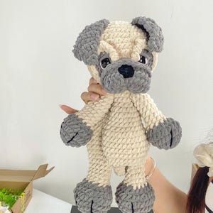 May include: A handmade crochet pug dog plushie in cream and gray. The pug has a cream-colored body with gray accents on the ears, paws, and snout. The plushie has black eyes and a black nose. The pug is a soft, cuddly toy.