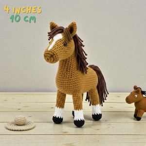 May include: A crocheted horse toy, approximately 4 inches (10 cm) tall, in shades of brown with a white blaze and black hooves. The horse has a dark brown mane and tail. A smaller version of the horse is also visible. A small, crocheted hat is in the foreground.
