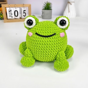 May include: A handmade, green crochet frog with large black eyes, pink cheeks, and a smiling mouth. The plush frog has a round body and four small legs. The frog is sitting on a white surface.
