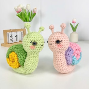 Könnte beinhalten: Zwei gehäkelte Schneckenspielzeuge. Eines ist hellgrün mit einem grün-gelben Gehäuse. Das andere ist rosa mit einem Regenbogengehäuse. Beide haben schwarze Augen, rosige Wangen und kleine Antennen. Die Schnecken sind handgefertigt und wirken weich.