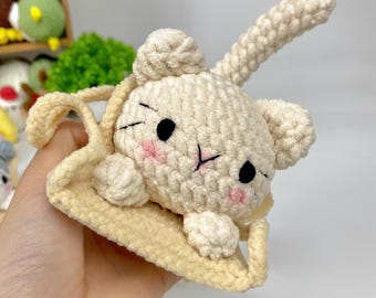 May include: A crocheted cream-colored cat plush toy with a long tail, sitting in a crocheted beige basket. The cat has black eyes and pink cheeks.