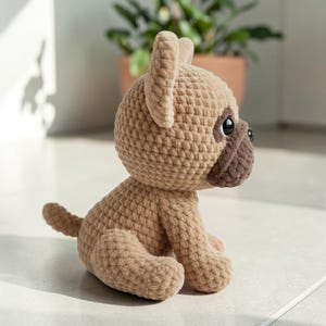 May include: A crocheted pug dog in a sitting position. The plush toy is tan with a darker brown muzzle and ears. The dog has black eyes and a small tail. The texture of the yarn is visible, giving the toy a handmade appearance.