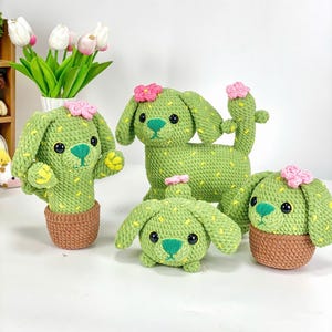 May include: Four crocheted cactus dog plushies in different sizes. The plushies are green with yellow accents and pink flowers. The plushies are sitting on a white surface.