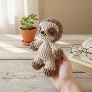 Puede incluir: Un juguete de perezoso tejido a ganchillo en tonos marrón y crema. El perezoso tiene los ojos cerrados y una sonrisa. Se sostiene en una mano, con una pequeña planta en maceta y un libro al fondo.