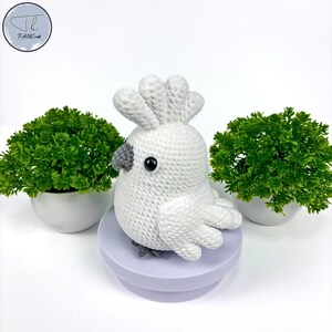 May include: White crocheted cockatoo bird with gray eyes and beak, sitting on a purple round base. The bird is surrounded by green potted plants.