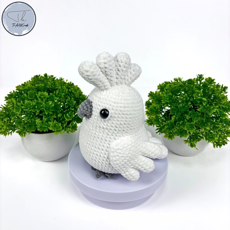 May include: White crocheted cockatoo bird with gray eyes and beak, sitting on a purple round base. The bird is surrounded by green potted plants.