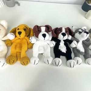 Può includere: Una collezione di sei cani di peluche all'uncinetto fatti a mano in vari colori. I cani includono crema, giallo, marrone e bianco, nero e bianco, grigio e bianco e beige e grigio. Ogni cane ha un design unico.