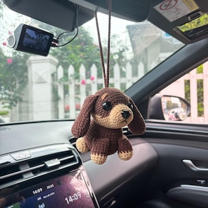Puede incluir: Un perro de crochet marrón colgando del espejo retrovisor de un coche. El perro tiene un cuerpo marrón, orejas beige y ojos marrones.