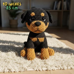 Op de afbeelding: Een handgemaakte gehaakte Rottweiler-puppy, zwart en bruin, zittend op een wit tapijt. De pluche hond is 25 cm hoog, met een gedetailleerd gezicht en slappe oren. De puppy heeft bruine markeringen op zijn gezicht, borst en poten.