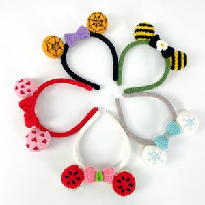 Può includere: Una collezione di fasce per capelli all'uncinetto colorate con vari disegni. Le fasce presentano disegni come una ragnatela, un'ape, cuori, fette di anguria e fiocchi di neve. Ogni fascia ha una combinazione di colori e un fiocco unici.
