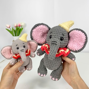 Może przedstawiać: Dwa szare szydełkowe słonie z różowymi i szarymi uszami, w żółtych kapeluszach i czerwono-białych kołnierzach. Słonie mają czarne oczy i różowe stopy. Jeden jest trzymany w dłoni osoby.