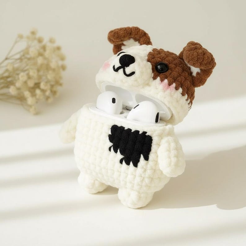 May include: A white and brown crocheted dog-shaped case for wireless earbuds. The case has a black patch on its chest and brown ears. The earbuds are white and visible inside the open case. The case is designed to protect and carry the earbuds.