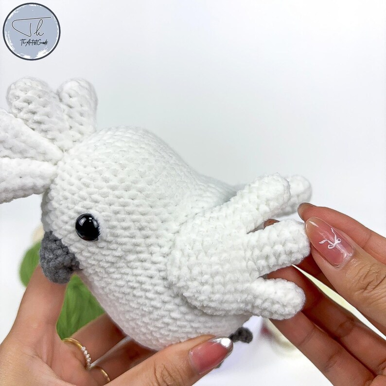 May include: A white crocheted cockatoo plush toy with a gray beak and black eyes. The toy is being held by a person's hands.