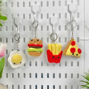 Op de afbeelding: Vier gebreide sleutelhangers met een voedselthema die aan haken hangen aan een wit prikbord. De sleutelhangers zijn een gebakken ei, een hamburger, frietjes en een pizzapunt. Elke sleutelhanger heeft een schattig cartoon gezicht.