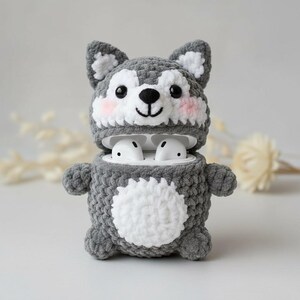 May include: A gray and white crocheted wolf-shaped case for wireless earbuds. The case has a white belly, pink cheeks, and black eyes. The earbuds are visible inside the open case. The case is designed to protect and carry the earbuds.