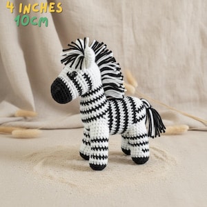 Puede incluir: Un juguete de cebra tejido a mano, con un cuerpo a rayas blanco y negro, una melena y una cola. El juguete mide aproximadamente 10 cm de altura. El texto "4 inches 10cm" es visible. La cebra está sobre una superficie arenosa.