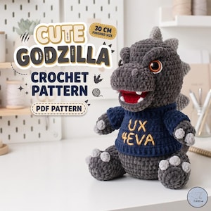 Op de afbeelding: Een donkergrijze gehaakte Godzilla knuffel met een marineblauwe trui met de gele tekst UX 4EVA. Het speelgoed heeft lichtgrijze klauwen en een rode mond met witte tanden. De tekst CUTE GODZILLA CROCHET PATTERN PDF PATTERN is zichtbaar.