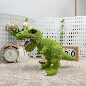 May include: A green crocheted Tyrannosaurus Rex dinosaur with white teeth and a pink mouth. The dinosaur has white accents on its feet and large, dark eyes. The toy is handmade and sits on a neutral-coloured surface.