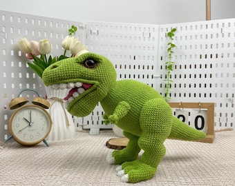 May include: A green crocheted Tyrannosaurus Rex dinosaur with white teeth and a pink mouth. The dinosaur has white accents on its feet and large, dark eyes. The toy is handmade and sits on a neutral-colored surface.