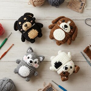May include: Four crocheted dog-shaped items in black, brown, gray, and white. Each has a unique design, with details like a white belly, pink cheeks, and black spots. The dogs are surrounded by yarn and crochet hooks, suggesting a handmade craft.