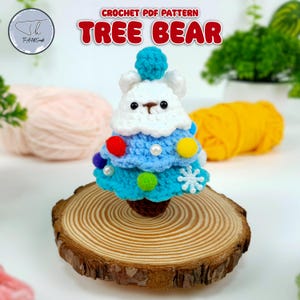 Könnte beinhalten: Häkelmuster für einen Baum-Bären, einen weißen Bären, der einen blauen und weißen Weihnachtsbaumhut mit bunten Ornamenten trägt. Der Bär sitzt auf einem Holzsockel.