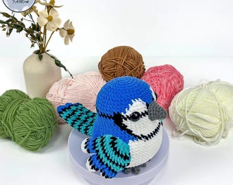 Puede incluir: Un pajarito azul tejido a crochet con el pecho blanco y alas negras y turquesa. El pájaro está sentado sobre una base morada y rodeado de ovillos de lana de varios colores.