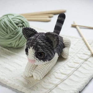 以下が含まれることがあります： 黒、白、グレーのハンドメイドかぎ針編み猫。 大きな表情豊かな目とピンクの鼻が特徴です。 猫は白いテクスチャードブランケットの上に横たわっており、背景には緑色の毛糸玉と木製の編み針があります。 魅力的で手作りのアイテムです。