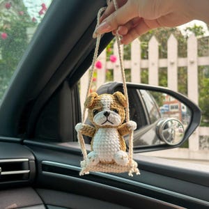 Puede incluir: Un cachorro de bulldog de crochet marrón y blanco colgando del espejo retrovisor de un coche. El cachorro está sentado en un pequeño columpio de crochet.