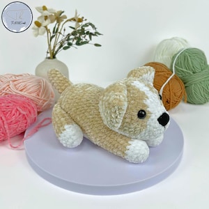 May include: A handmade crochet dog in tan and white, lying on a light purple surface. The dog has black eyes and a black nose. Balls of yarn in pink, white, green, and brown are in the background, along with a small vase of flowers.