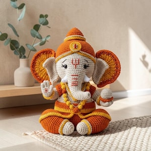 Könnte beinhalten: Gehäkelte Ganesh-Figur in sitzender Position. Die elefantenköpfige Gottheit ist hauptsächlich cremefarben, orange und gelb. Die Figur hat große Ohren, einen Rüssel und ist mit einer Krone und Schmuck geschmückt. Die Figur sitzt auf einem kleinen Teppich.