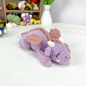 Könnte beinhalten: Ein handgehäkelter Drache in Lavendel- und Mauve-Tönen. Das Plüschtier hat detaillierte Schuppen, Flügel und kleine weiße Hörner. Der Drache liegt und zeigt sein weiches, strukturiertes Design. Weitere Häkeltiere sind im Hintergrund zu sehen.