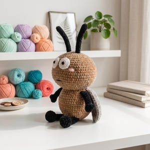 May include: A plush toy shaped like a bug with a round tan body, black limbs, and two black antennae. It features large white eyes with black pupils and small pink blush marks on its cheeks. The toy has a textured, soft velvet-like appearance.