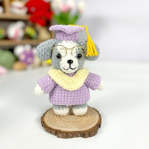 Op de afbeelding: Gehaakte hondenfiguur met een afstudeerhoed en bril. De hond is grijs en wit met een paarse hoed en trui, en een gele kwast. Hij staat op een houten schijf.