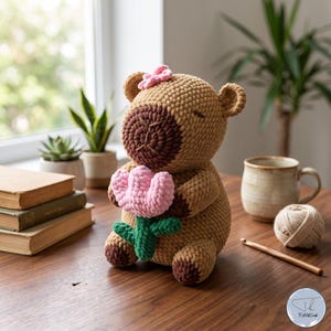 May include: A crocheted capybara plush toy in light brown yarn with dark brown paws and snout. The capybara has a small pink flower on its head and is holding a pink and green crocheted tulip flower. The toy features closed eyes and rounded ears.