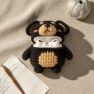 May include: A black and tan crochet bear-shaped case for wireless earbuds. The case has a tan muzzle and belly, with pink blush details. The earbuds are white. A pencil and paper are in the foreground, and a ball of yarn is in the background.