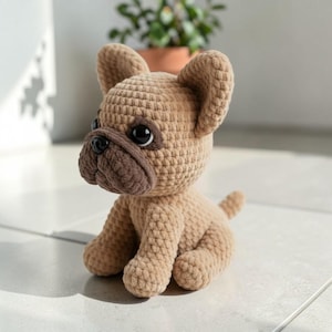 May include: A crocheted French Bulldog dog in a sitting position. The plush toy is tan with a darker brown muzzle and ears. It has large, black eyes and a small tail. The dog is handmade and sits on a light-colored surface.