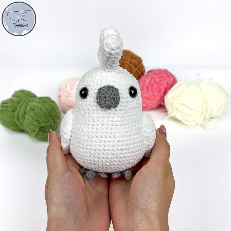 May include: White crocheted cockatoo bird with black eyes and a gray beak. The bird is sitting in the palms of a person's hands. There are balls of yarn in the background.
