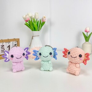 May include: Three crocheted axolotl keyrings in pastel shades: pink, mint green, and peach. Each axolotl features a smiling face, black eyes, and frilly gills in contrasting colours. The keyrings are displayed on a white surface with decorative flowers.
