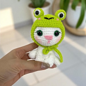 May include: A handmade white crochet doll wearing a green frog hat with large black eyes and a pink nose. The doll has a green scarf and is held in a person's hand. The background is blurred, with green plants visible.