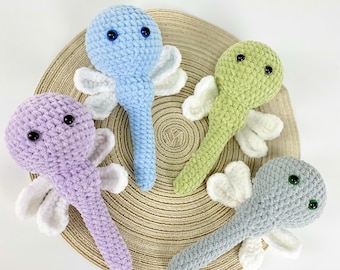May include: Four handmade crochet dragonfly toys in pastel colors: lavender, blue, green, and gray. Each dragonfly has white wings and black or blue eyes. The toys are arranged on a round, woven mat.