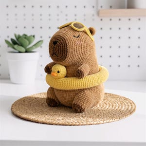 Peut inclure: Un jouet en peluche capybara crocheté marron portant des lunettes de natation jaunes et une bouée jaune autour de la taille. Le capybara tient un petit caneton crocheté jaune. Il présente des yeux fermés brodés et des joues roses.
