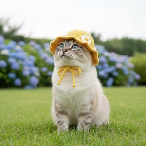 May include: A cat wearing a yellow crocheted hat with a white flower and a matching bow. The cat has blue eyes and is sitting on green grass with blue flowers in the background.