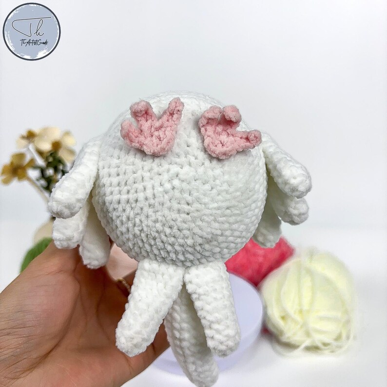 May include: White crocheted bird amigurumi with pink felt wings. The bird has a round body and long legs.